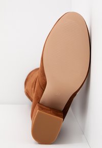 Bottine en daim marron avec semelle lisse striée et talon bloc, taille 37, positionnée debout contre un coin blanc en arrière-plan.