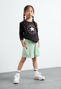 Crna majica dugih rukava s grafikom "Converse All Star", mentol zelene šortse s džepovima i bijele tenisice visoke s crnim akcentima.