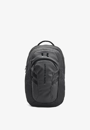 Sac à dos noir en matériau durable avec un dos rembourré, plusieurs compartiments, des fermetures éclair latérales et un logo proéminent à l'avant.