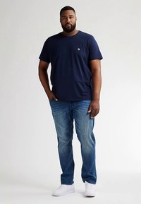 Tmavě modré bavlněné tričko s kulatým výstřihem a malým logem, kombinované s světle modrými džínami a bílými teniskami. Model stojí vzpřímeně.