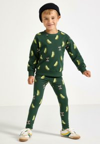 Grøn bomulds sweatshirtsæt og leggings medært-bælg mønster og teksten "Peas & Love", parret med en sort strikket hat og sneakers.