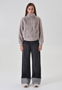 Giacca in finta pelliccia con zip di colore grigio chiaro, con colletto alto e polsini elasticizzati. Abbinata a jeans scuri a gamba larga con vita alta e scarpe bianche.