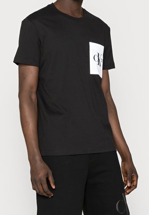 T-shirt in cotone nero con maniche corte, caratterizzato da una tasca bianca con un logo. Forma semplice e aderente, texture liscia.