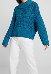 Pull tricoté couleur sarcelle avec un col montant, présentant un motif texturé et un ourlet et des poignets côtelés. Associé à un pantalon blanc.