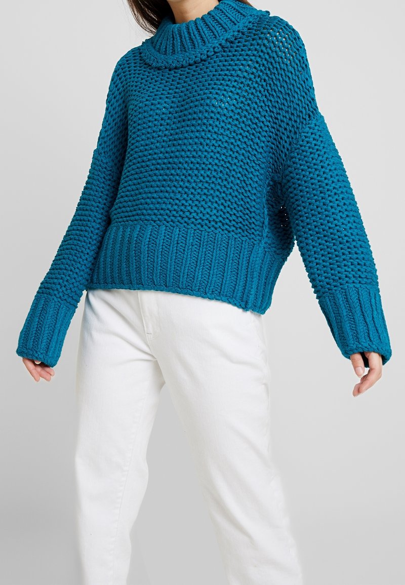 Pull tricoté couleur sarcelle avec un col montant, présentant un motif texturé et un ourlet et des poignets côtelés. Associé à un pantalon blanc.