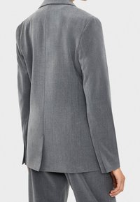 Person klädd i en grå skräddarsydd kavaj och matchande byxor, sedd bakifrån mot en enkel ljus bakgrund.