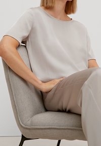 Top short a maniche corte color beige chiaro realizzato in tessuto morbido, abbinato a pantaloni coordinati, seduto su una sedia grigia testurizzata. Design minimalista.