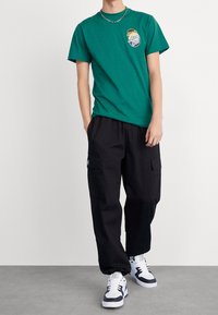 Vihreä puuvillainen t-paita, jossa on rintalogo, yhdistetty mustiin taskurulliin sivutaskuilla ja valkoisiin lenkkareihin mustilla yksityiskohdilla.