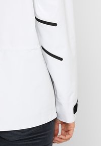 Chemise blanche affichant une finition lisse et brillante avec des accents noirs le long des manches et un détail de poignet en velcro.