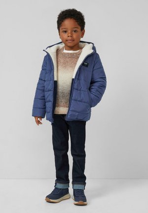 Blauwe pufferjack met een witte fleece-gevoerde capuchon, gecombineerd met een kleurige gebreide trui, donkere jeans en blauwe sneakers.