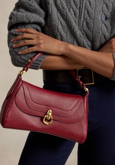 Persona que lleva un suéter gris de punto trenzado y pantalones oscuros, sosteniendo un pequeño bolso de cuero rojo con un broche dorado.