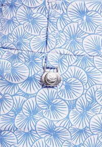 Patterned fabric in light blue and white features circular floral motifs. A silver metal button with a heart design fastens a pocket.