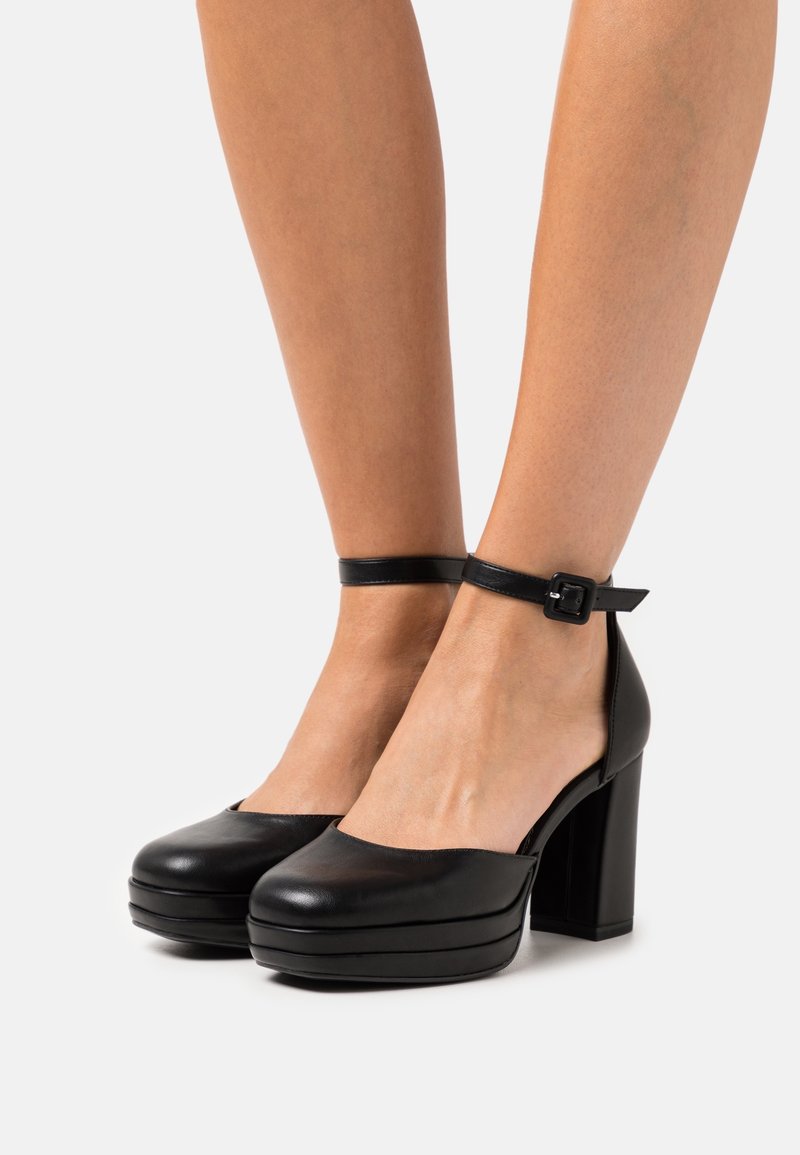 Jambes portant des talons compensés en cuir noir avec des brides à la cheville et des talons carrés sur un fond blanc uni.