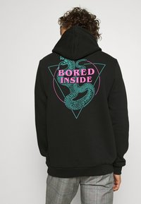Persona di spalle che indossa una felpa nera con design di serpente turchese e testo rosa "BORED INSIDE", abbinata a pantaloni a quadri grigi.