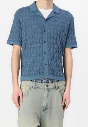 Niebieska koszula z krótkim rękawem o fakturowanym zygzakowatym wzorze, z kołnierzykiem i zapięciem na guziki, zestawiona z wyblakłymi jasnymi dżinsami.