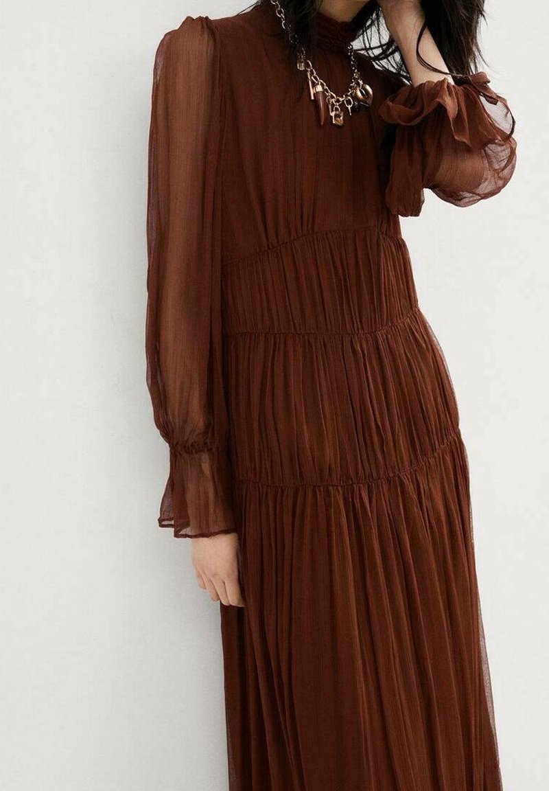 Femme portant une robe marron transparente à manches longues et col montant, avec des plis étagés et un collier chaine doré épais.