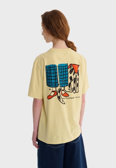 Personne aux cheveux rouges attachés porte un T-shirt jaune représentant des jambes de dessin animé en pantalon à carreaux bleus, des chaussures rouges et un chien tacheté avec un bandana rouge.