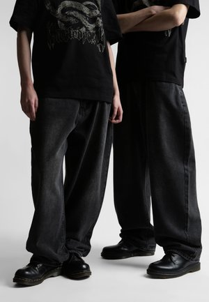 Dwie osoby stojące obok siebie w czarnych koszulkach z nadrukiem, luźnych ciemnych dżinsach i czarnych skórzanych butach na jednolitym tle.