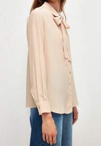 Blusa realizzata in un tessuto leggero e morbido di color pesca chiaro, presenta un colletto, un fiocco al collo e bottoni dorati sul davanti e ai polsini.