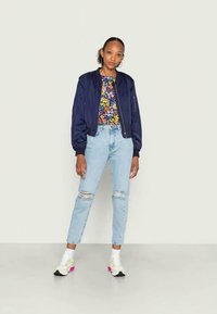 Jovem mulher de pé, vestindo uma jaqueta bomber azul marinho, camisa floral, jeans claros rasgados e ténis brancos, contra um fundo claro e liso.