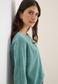 Teal kordfloddsweater med rund halsringning och ribbad struktur, med långa ärmar och en avslappnad passform, fotograferad mot en neutral bakgrund.