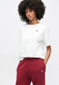 Νεαρή γυναίκα με μπούκλες, που φοράει λευκό μπλουζάκι και μπορντό παντελόνια, και τα δύο με μικρά μαύρα αθλητικά λογότυπα, στέκεται με τα χέρια στις τσέπες.