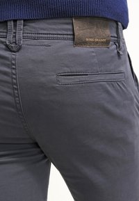 Donkergrijze broek met een gladde textuur, voorzien van een achterzak en een leren label met de tekst "BOSS ORANGE" aan de taille.