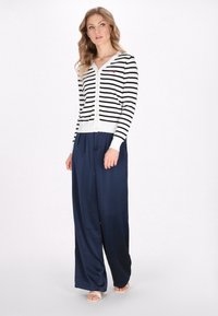 Cardigan striped în alb și negru cu decolteu în V și închidere cu nasturi, asociat cu pantaloni largi navy și sandale deschise.