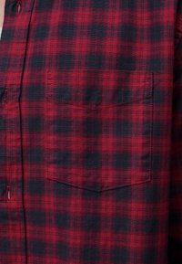 Πουκάμισο φανέλα με κόκκινο και μαύρο καρό, με ορατή τσέπη στο στήθος και κουμπότρυπες κατά μήκος της μπροστινής αριστερής πλευράς.