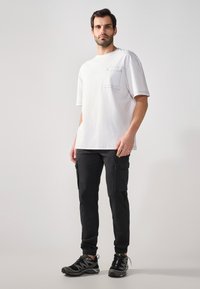 T-shirt bianco in jersey con vestibilità ampia e taschino sul petto, abbinato a pantaloni cargo neri con tasche laterali. Scarpe da ginnastica nere completano il look.