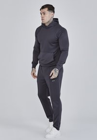 Sötét szürke kapucnis pulóver és hozzá illő melegítőnadrág puha anyagból, elülső zsebbel, bordás mandzsettákkal és fehér sportcipővel. Minimalista dizájn.