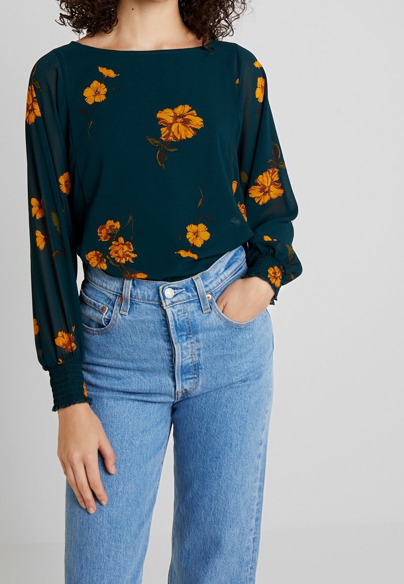 Femme portant un chemisier à manches longues turquoise avec un imprimé floral orange, rentré dans un jean bleu clair taille haute, une main dans la poche.