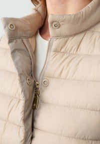 Giacca puffer beige chiaro con chiusura zip, colletto con bottoni a pressione e texture trapuntata. Caratterizzata da dettagli in oro e tessuto morbido.
