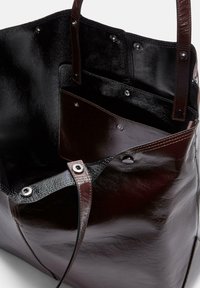 Brązowa skórzana torba tote o gładkiej fakturze, z wieloma wewnętrznymi kieszeniami i srebrnymi akcentami. Otwarta góra z krótkimi uchwytami.