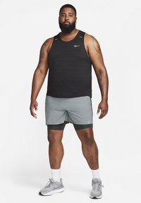 Črna Nike majica brez rokavov s teksturiranim dizajnom, sivi Nike šorts in sivi športni čevlji. Ima okroglo ovratnico in ramenske poudarke.
