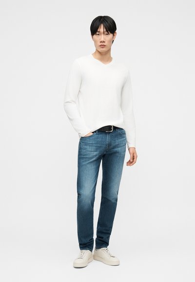 Νεαρός άνδρας στέκεται φορώντας λευκό πουκάμισο με μακριά μανίκια, μπλε τζιν με μαύρη ζώνη και λευκά αθλητικά παπούτσια, μπροστά σε απλό ανοιχτόχρωμο φόντο.