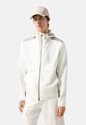 Jonge man met een off-white zip-up jas met capuchon en beige pet, staand met een neutrale uitdrukking tegen een eenvoudige achtergrond.