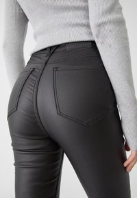 Zwarte faux leren broek met een aansluitend ontwerp, voorzien van twee achterzakken en zichtbare stiksels, gecombineerd met een lichtgrijze ribbeltrui.