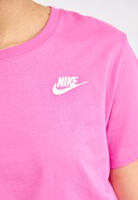 Rožinė medvilninė marškinė su apvalia iškirpte, kurių kairėje krūtinės pusėje yra baltas siuvinėjimas Nike logotipas. Lygus paviršius.