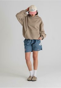 Beige overdimensjonert hettegenser med en frontlomme, kombinert med blå shorts med hvite striper og en logo. Hvite sokker og lysegrå sko fullfører ensemblet.