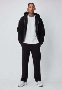 Jeune homme tatoué portant un sweat à capuche noir zippé, un t-shirt blanc, un pantalon noir et des baskets blanches, debout devant un fond uni.