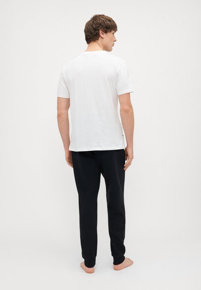 Balta kokvilnas t-krekls ar īsām piedurknēm un apaļu kakla izgriezumu, kombinēts ar melnajām sporta biksēm ar ribotām aprocēm, redzams no aizmugures.