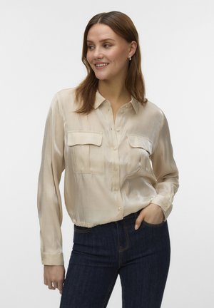 Lachende vrouw die een glanzend beige blouse met knopen en twee borstzakken draagt, samen met donkerblauwe jeans, staand met één hand in de zak.