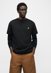 Άνδρας που φοράει μαύρο μπλουζάκι μακρυμάνικο κάτω από μαύρο κοντομάνικο μπλουζάκι με μικρό κίτρινο λογότυπο και καφέ παντελόνι, με τα χέρια στις τσέπες, έκφραση ουδέτερη.