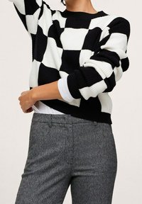 Zwart-witte damessweater met ruitjespatroon en geribbelde textuur. Heeft een ronde hals en lange mouwen. Gedragen over een wit shirt en grijze broek.