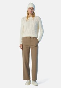 Donna con un berretto di maglia bianco, un maglione crema a treccia, pantaloni dritti beige e sneakers bianche, in piedi con una mano in tasca.