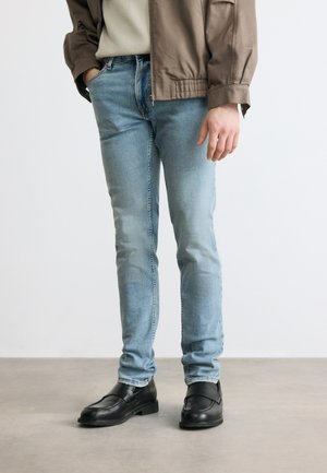 Blugi din denim albastru deschis, croi strâmt, purtați cu mocasini negri din piele și o jachetă maro, persoană stând pe un podea netedă, în fața unui perete alb.