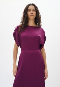 Robe violette en tissu texturé, avec des manches larges et une taille cintrée. Finition lisse avec un décolleté bateau et un design long jusqu'au sol.