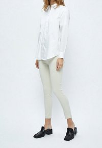 Valkoinen pitkähihaisena nappipaita etusululla, yhdistettynä vaaleanbeigeihin vartalonmyötäisiin housuihin ja mustiin muloihin, joissa on neliskulmainen kärki ja paksu korko.