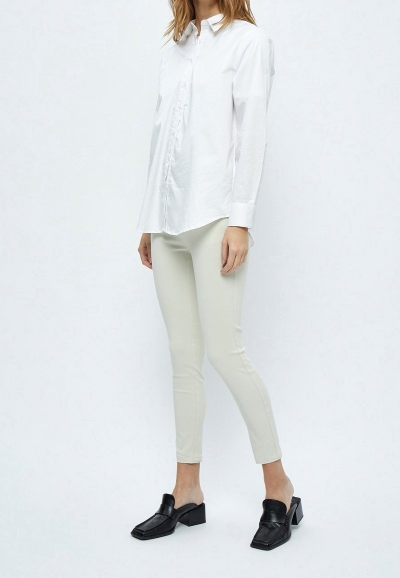 Valkoinen pitkähihaisena nappipaita etusululla, yhdistettynä vaaleanbeigeihin vartalonmyötäisiin housuihin ja mustiin muloihin, joissa on neliskulmainen kärki ja paksu korko.